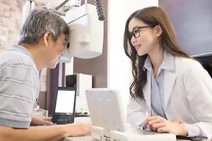 A man and a woman are sitting at a desk and the woman is wearing glasses and the man is looking at her through a device