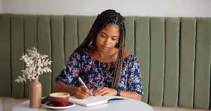 A woman is sitting on a chair in front of a table in a cafe, writing in a book with a pen. She is wearing a blue blouse and has braided hair. There is a cup of coffee, a saucer, and a potted plant on the table. Behind her, there is a green wall with a partition. The light is shining on her face.