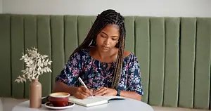 A woman is sitting on a chair in front of a table in a cafe, writing in a book with a pen. She is wearing a blue blouse and has braided hair. There is a cup of coffee, a saucer, and a potted plant on the table. Behind her, there is a green wall with a partition. The light is shining on her face.