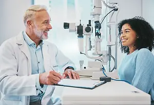 An elderly male doctor in a white coat sits beside a female patient in a blue sweater as they converse while the doctor holds a pen and paper in front of an ophthalmoscope machine
