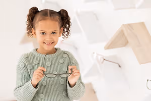 A smiling young girl holding a pair of glasses in front of a white wall with wooden shelves and a wooden structure