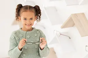 A smiling young girl holding a pair of glasses in front of a white wall with wooden shelves and a wooden structure