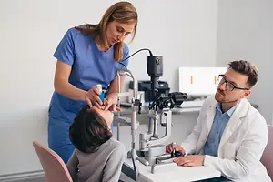 A man wearing glasses is examining a child's eyes with a machine while a woman in blue scrubs is holding a bottle.