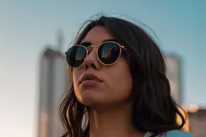 A woman wearing sunglasses is looking up at the sky with a city skyline in the background.