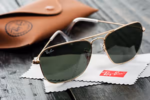 Ray-Ban sunglasses with brown frame and green lenses on a white cloth with a brown leather case in the background.