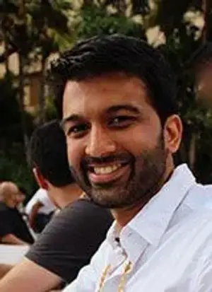 A smiling man wearing a white polo shirt with a gold necklace and a black-haired man wearing a black shirt behind him.