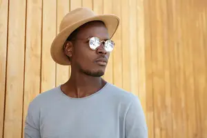 An adult man with a beard wearing a hat and sunglasses is standing against a wooden wall.