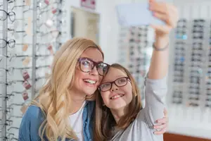 A woman and a young girl wearing glasses are standing close to each other and smiling in a store while taking a selfie