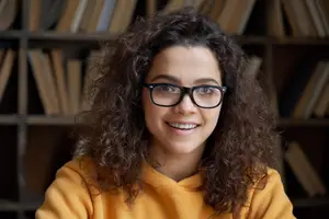 Portrait of a woman with curly hair, wearing glasses, sitting in a library, and smiling at the camera.
