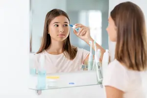 A woman is applying eye drops in front of a mirror, while another woman is watching her from behind.
