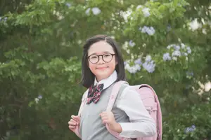 A young girl with glasses and a school uniform is smiling and looking forward, carrying a pink backpack, standing in front of a blurry background of green trees and plants with white flowers.