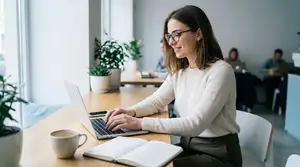 A woman works on a laptop in an office with other people in the background.