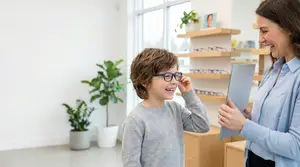 Young boy wearing glasses, standing in front of his mother at a optometrist's office