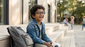 A young boy wearing glasses and a denim jacket sitting on steps with a backpack and holding a soccer ball.
