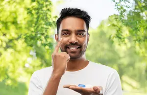 A man in a white t-shirt is pointing at his face and smiling while holding a contact lens.