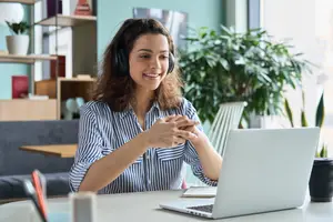 A smiling woman wearing headphones and a striped shirt is sitting at a desk with a laptop in front of her.