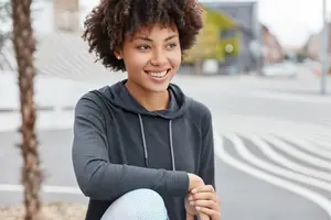 A woman with curly hair is smiling and posing for a photo in an outdoor area. She is wearing a black hoodie and white leggings. Behind her is a blurry view of a street with a tree and a building with a yellow wall. The road has white markings and a pole. There are also blurry cars driving on the road.