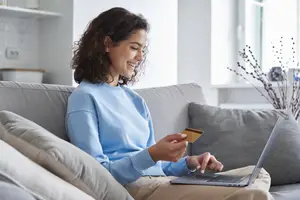 A woman sitting on a couch is holding a credit card and a laptop in her hands