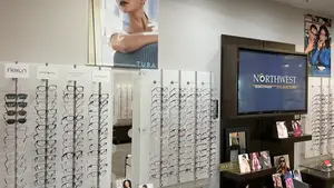 A shop interior with display boards, a monitor, shelves, and a desk. Eyeglasses are on the wall, and there are chairs and a desk with a monitor on top.