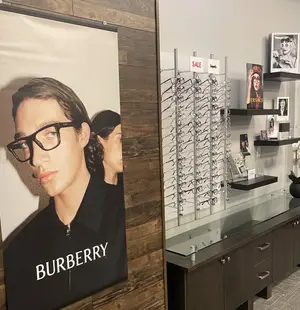 Inside a Burberry store, a wooden wall has a poster of a man wearing glasses. Behind the poster is a desk with a glass top and various items on display, including glasses and a vase with flowers. The glass top has several glasses on display. The store has a modern design, with wooden walls and a glass display case.