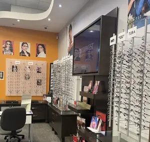 Interior of a retail store specializing in eyewear with displays of frames and a large monitor showing a woman wearing glasses, accompanied by a desk, chairs, and other office furniture.
