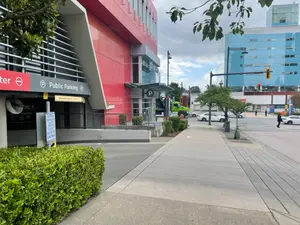 A public parking area with a red building, a sidewalk, and street signs, with a person walking on the sidewalk and a green truck parked nearby.