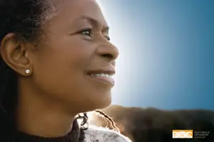 A woman with curly hair is smiling and looking towards the sun with her eyes closed.