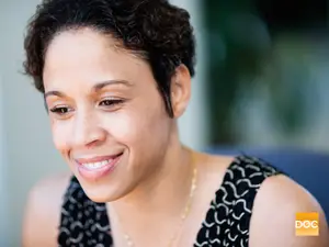 Woman with short curly hair wearing a black and white patterned sleeveless top, smiling at the camera with her eyes closed