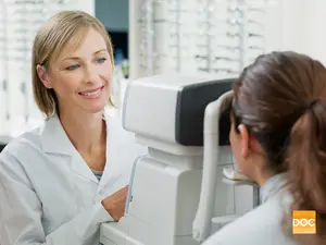 A woman with blonde hair in a white lab coat is smiling at another woman in a white lab coat who is looking at her through an eye exam machine.