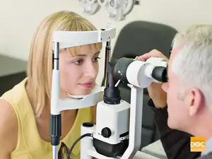 A woman in a yellow shirt is being examined by a doctor with a white and black ophthalmoscope.