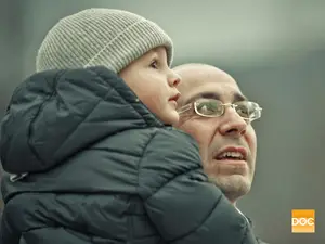 A man in glasses holds a child wearing a beanie and a black jacket, smiling while looking up.