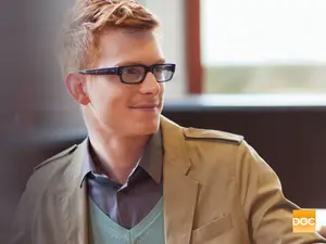 A man wearing glasses and a beige jacket is sitting inside a room with a window behind him, smiling and looking to the right. The text 'DOC' is visible in the bottom right corner.
