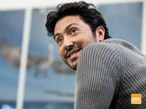 A man with a beard and mustache wearing a gray sweater looks out the window with a smile on his face