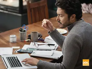A man eats cheese while working on a laptop at a desk.