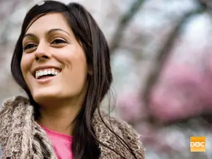 Portrait of a smiling woman with long black hair and a pink shirt, standing in front of a tree with pink flowers, possibly in a park, with a watermark in the bottom right corner