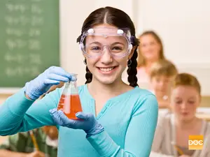 A young girl is conducting a science experiment in a classroom, wearing protective goggles, gloves, and holding a container with a red liquid.