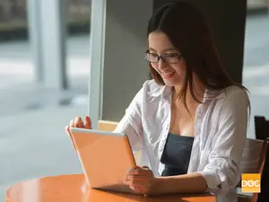 A smiling woman wearing glasses is sitting on a chair and holding a tablet while looking at the screen.