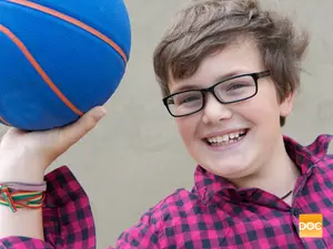 A smiling boy with glasses holds a basketball, showing a cheerful expression and wearing a plaid shirt and bracelets.
