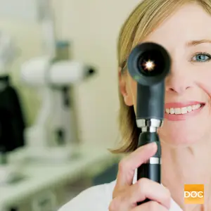 A woman doctor with a stethoscope around her neck smiles and looks into a microscope.