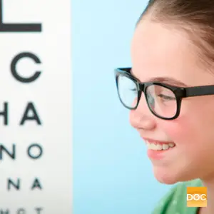 A girl wearing glasses is smiling and looking at a blurry eye chart with the letters CHA and DOC on it.