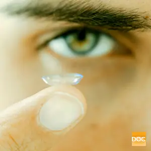 A closeup of a man holding a contact lens in his finger, with his eye in the background and a watermark with the text DOC in the bottom right corner.