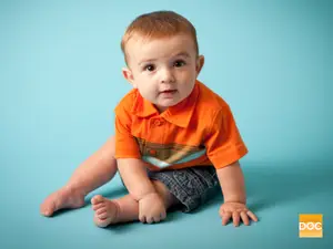 A baby boy with short hair and wearing an orange polo shirt is sitting on a blue background and looking to his left.