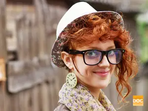 A woman with red curly hair wearing a floral hat and glasses, with a vintage clock earring, smiling at the camera