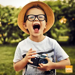 A young boy wearing a straw hat, black glasses, and a t-shirt is holding a camera and posing for a photo in a grassy area.
