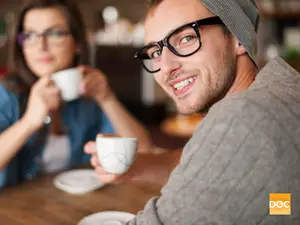 A man wearing glasses and a woman wearing a blue shirt sitting at a table in a cafe drinking coffee