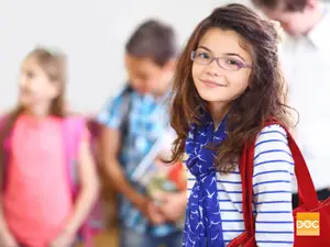 A young girl with glasses and a scarf is smiling and posing for a picture with other children in the background.