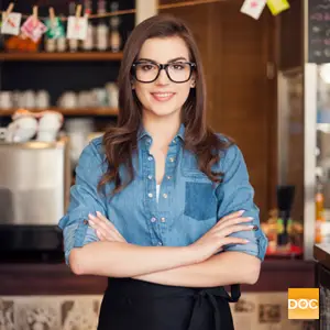 A woman wearing glasses and a denim shirt is smiling and posing for a photo in a coffee shop