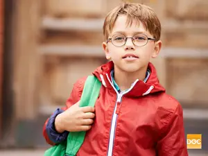 A boy with a red jacket and glasses is holding a green backpack