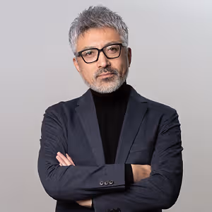 A man with gray hair, a beard, and glasses stands with his arms crossed in a studio setting against a gray background.