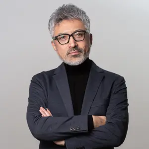 A man with gray hair, a beard, and glasses stands with his arms crossed in a studio setting against a gray background.